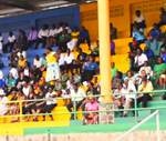 Crowd seated in colorful stadium bleachers during an outdoor funeral service.