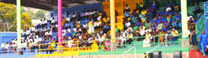 Crowd seated in colorful stadium bleachers during an outdoor funeral service.