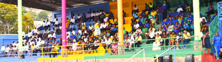 Crowd seated in colorful stadium bleachers during an outdoor funeral service.