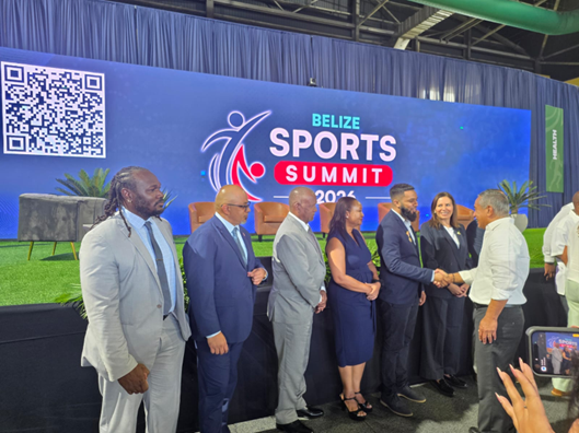 Delegates and officials shaking hands on stage in front of a Belize Sports Summit backdrop.